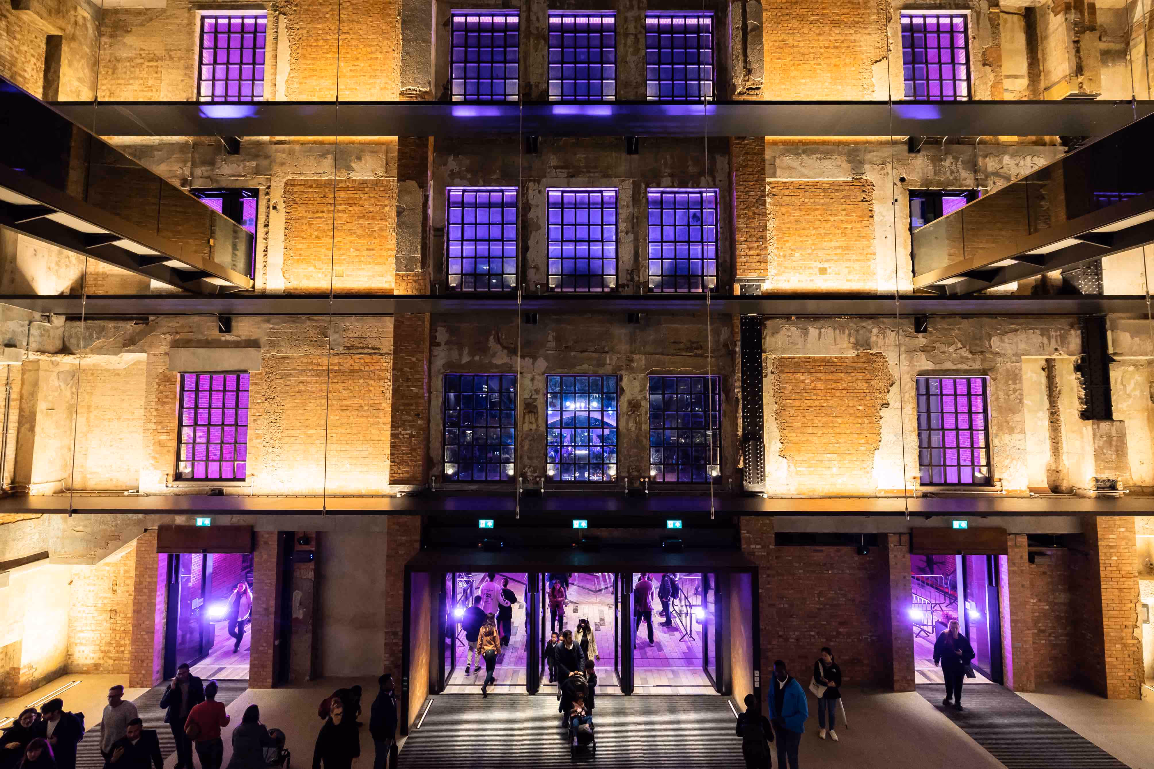 Inside Battersea Power Station, crowds enter through the door into the multi-storey foyer; the large windows show the purple lights from outside.