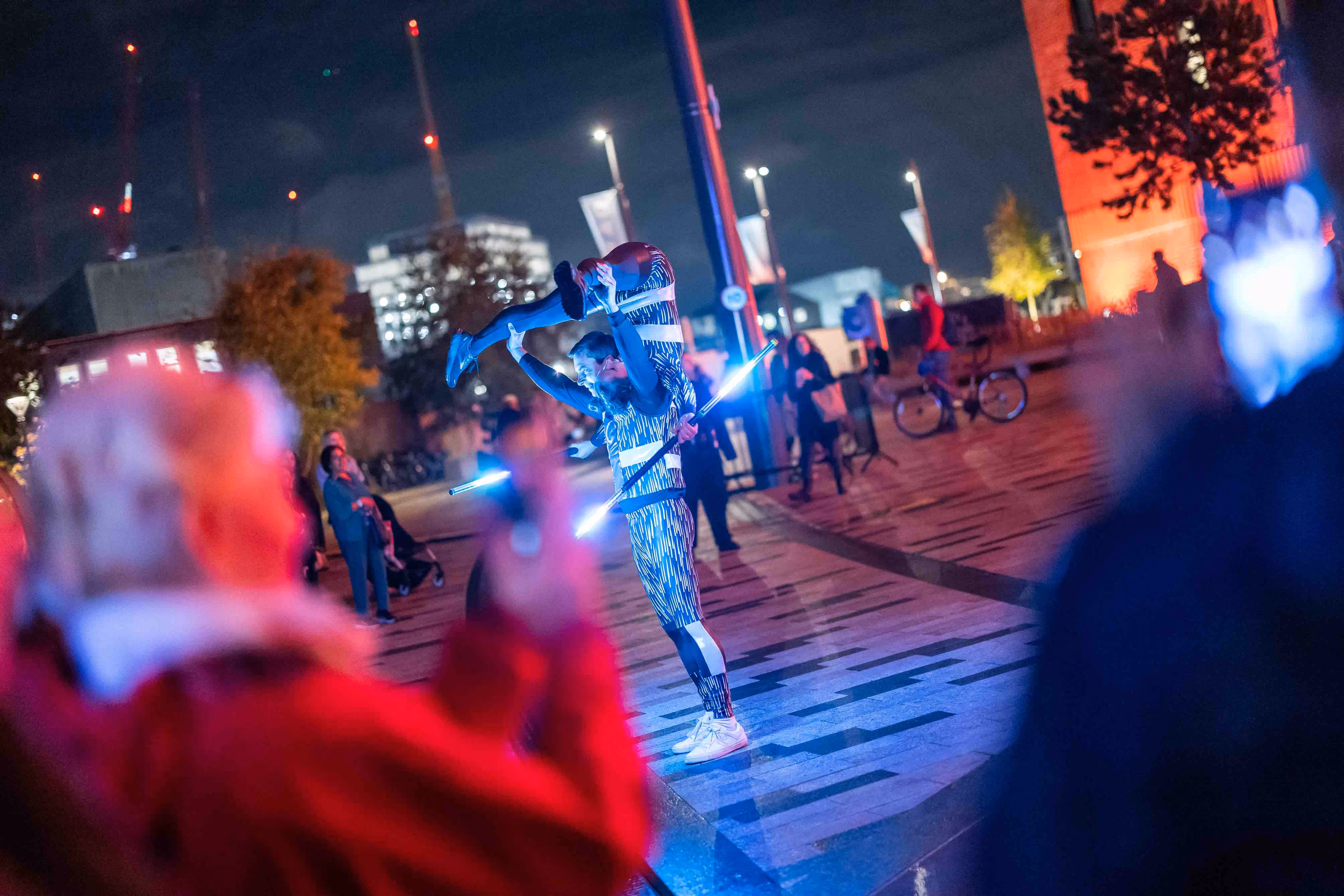 Street performers with large rods that are lit on each end perform acrobatics outside the entrance of Battersea Power Station whilst the crowd watches.