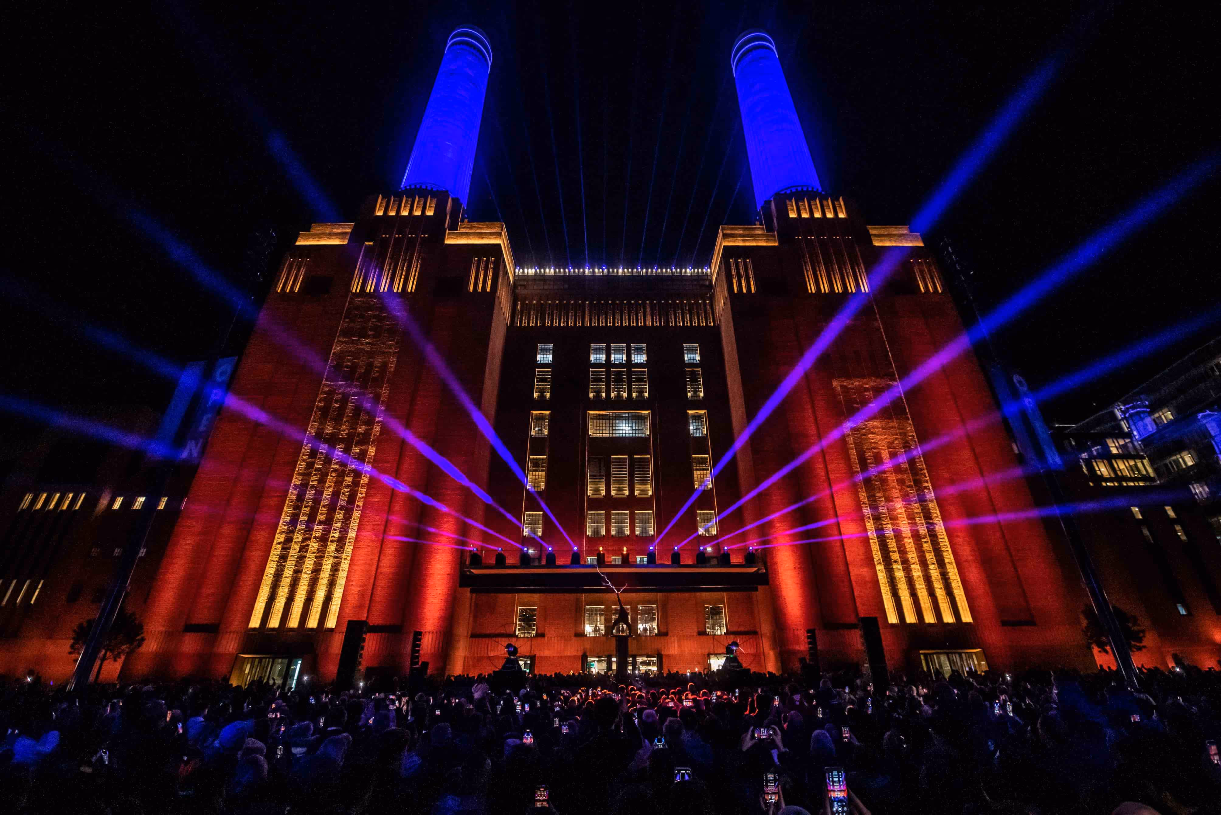 Battersea Power Station launch event; the station at night, uplit with red and blue lights with a large crowd in front of the building.