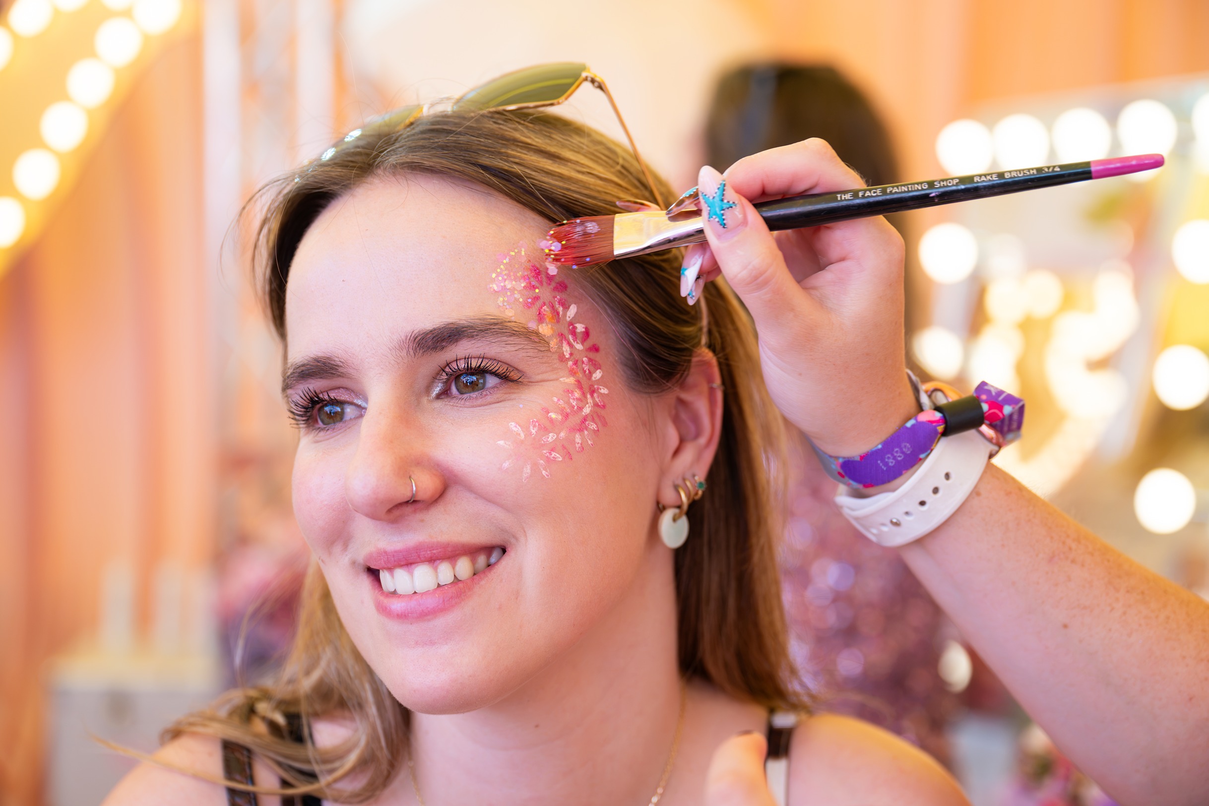 Festival-goer having face paints and glitter put on her face at 'Backstage with Dove' beauty activation by Bearded Kitten