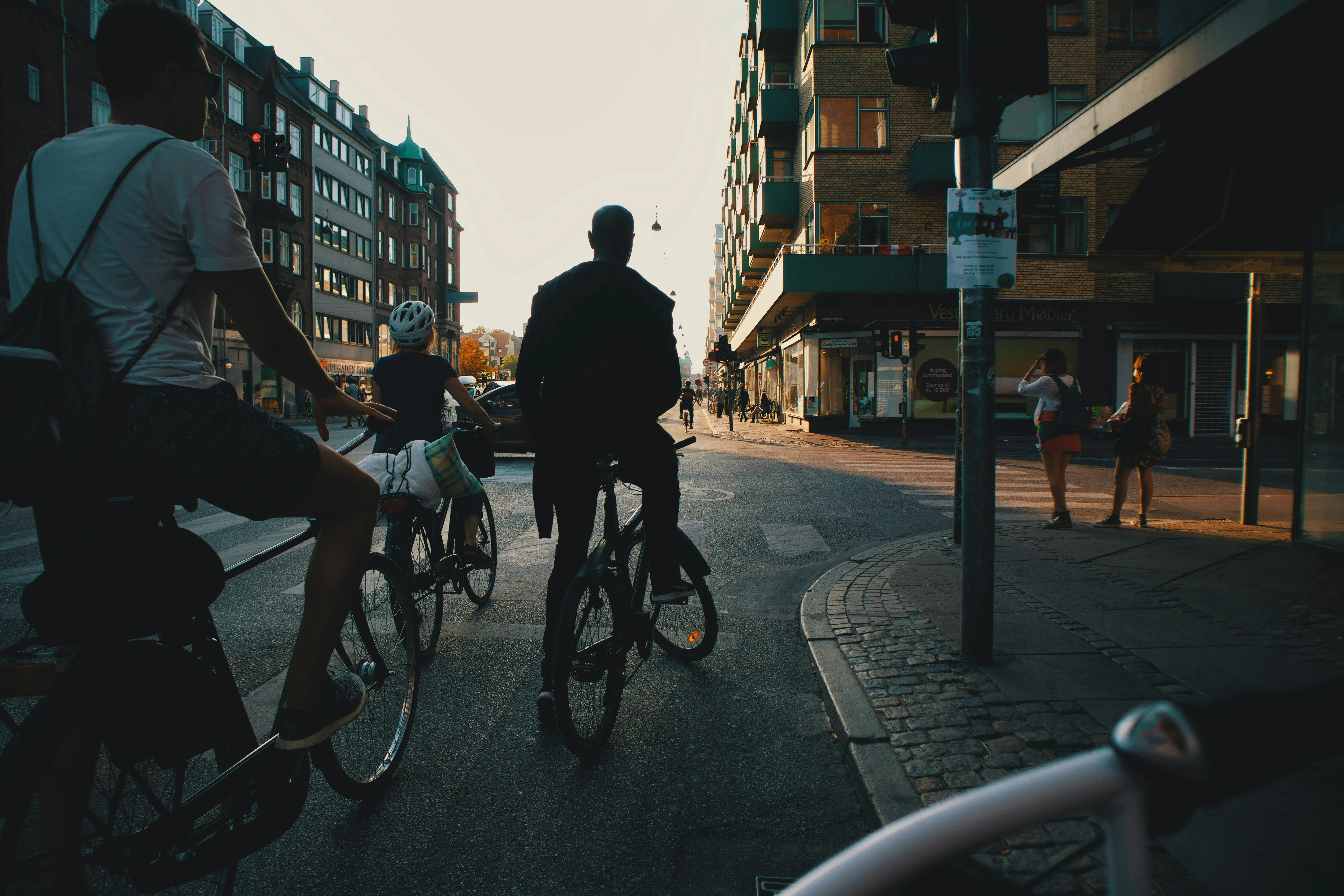 Cyclists waiting at a city junction during golden hour, with buildings, pedestrians and warm evening light illuminating the street.