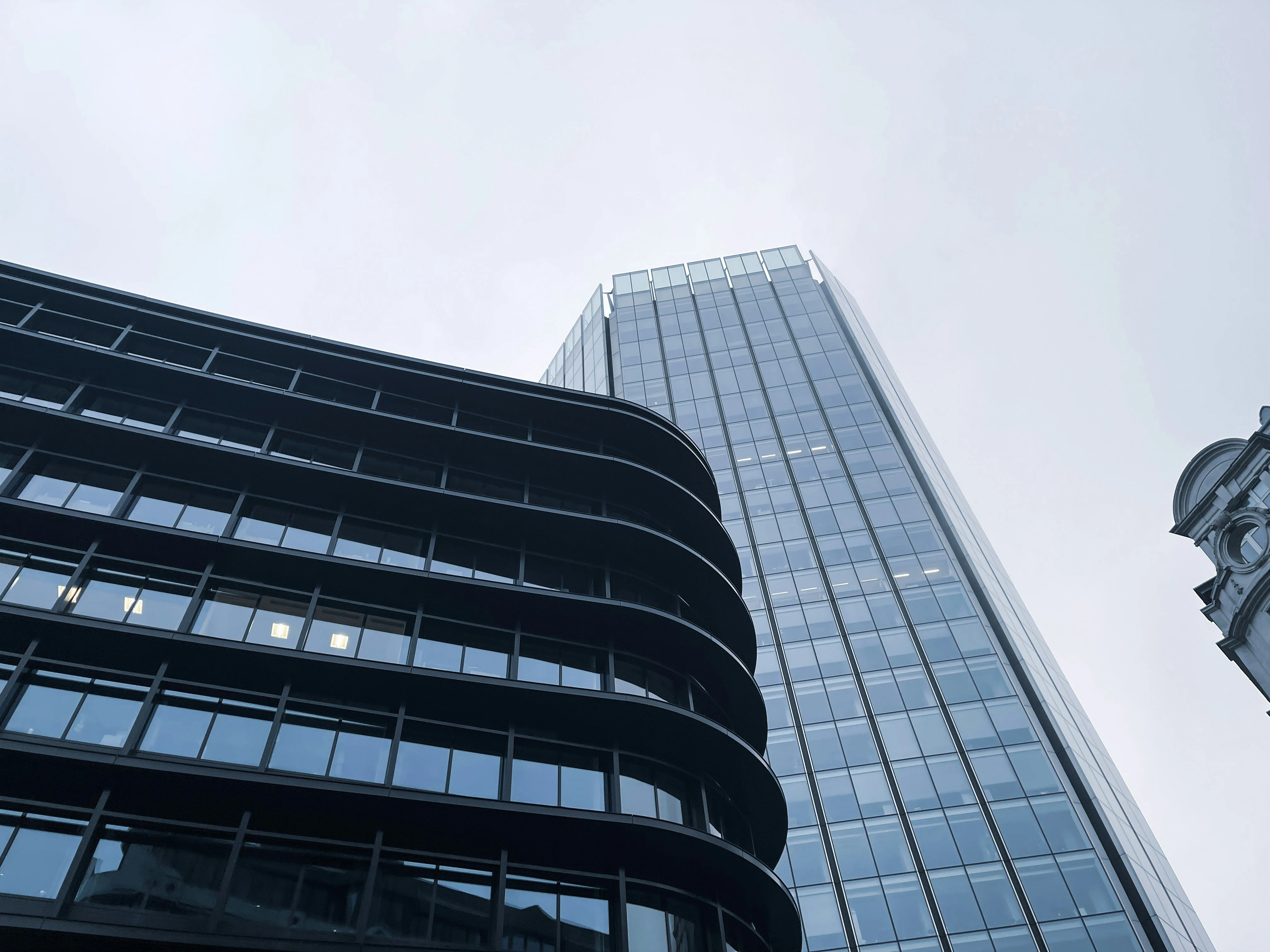 Upward view of modern glass office buildings against an overcast sky, featuring a curved dark façade in the foreground and a tall reflective tower behind it.
