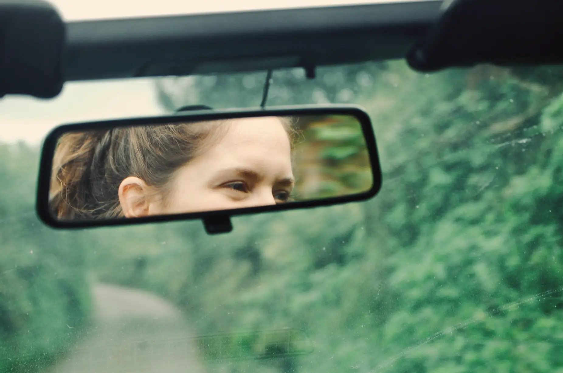 Close-up view through a car’s windshield, showing a rear-view mirror reflecting the eyes and upper face of a person driving. The background outside the car is blurred greenery, giving a sense of motion or a rural road.