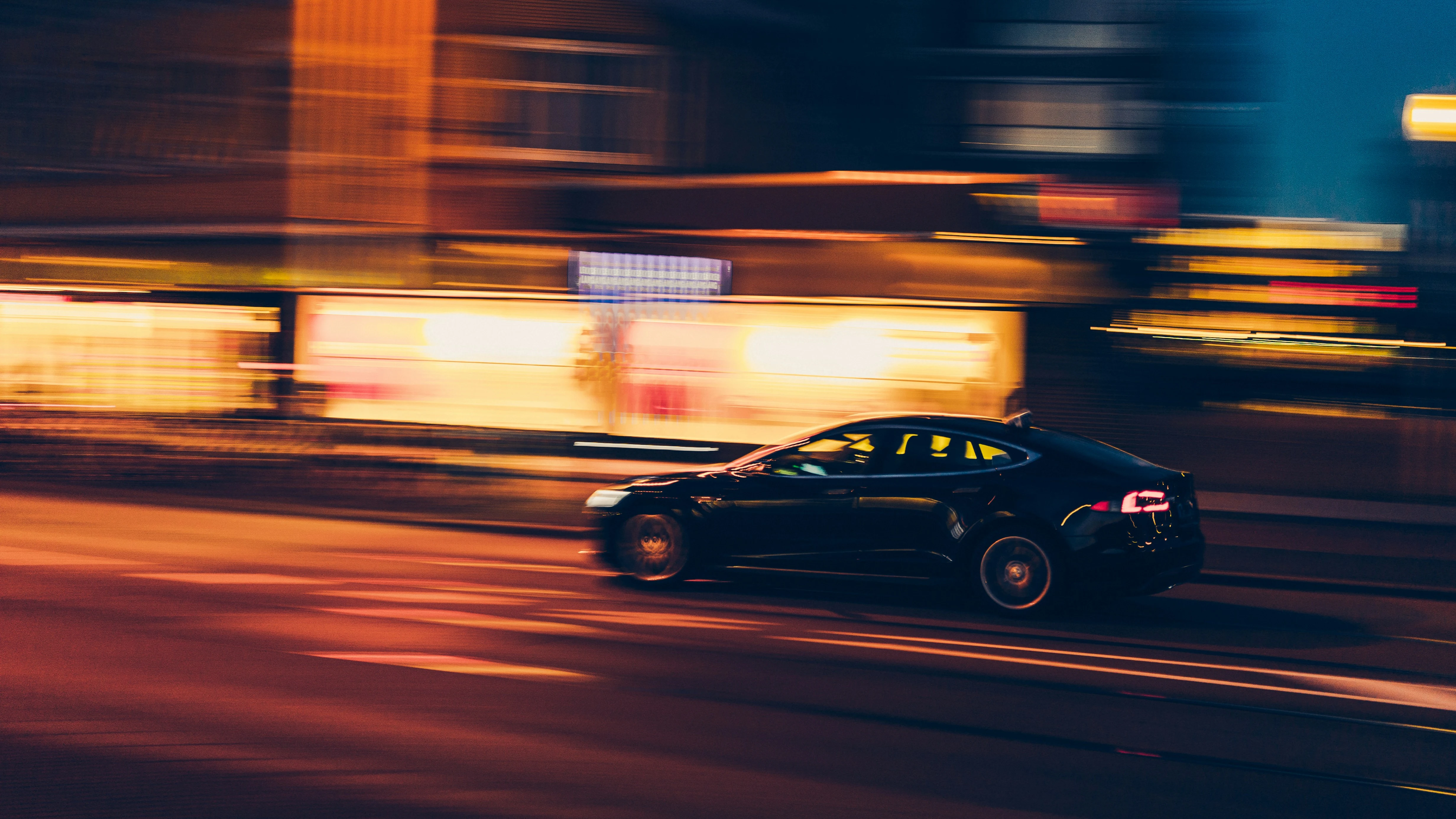 A black Tesla sedan speeding through a city street at night, captured with a motion-blur effect. Bright shopfront lights and neon colours streak across the background, emphasizing the sense of speed.