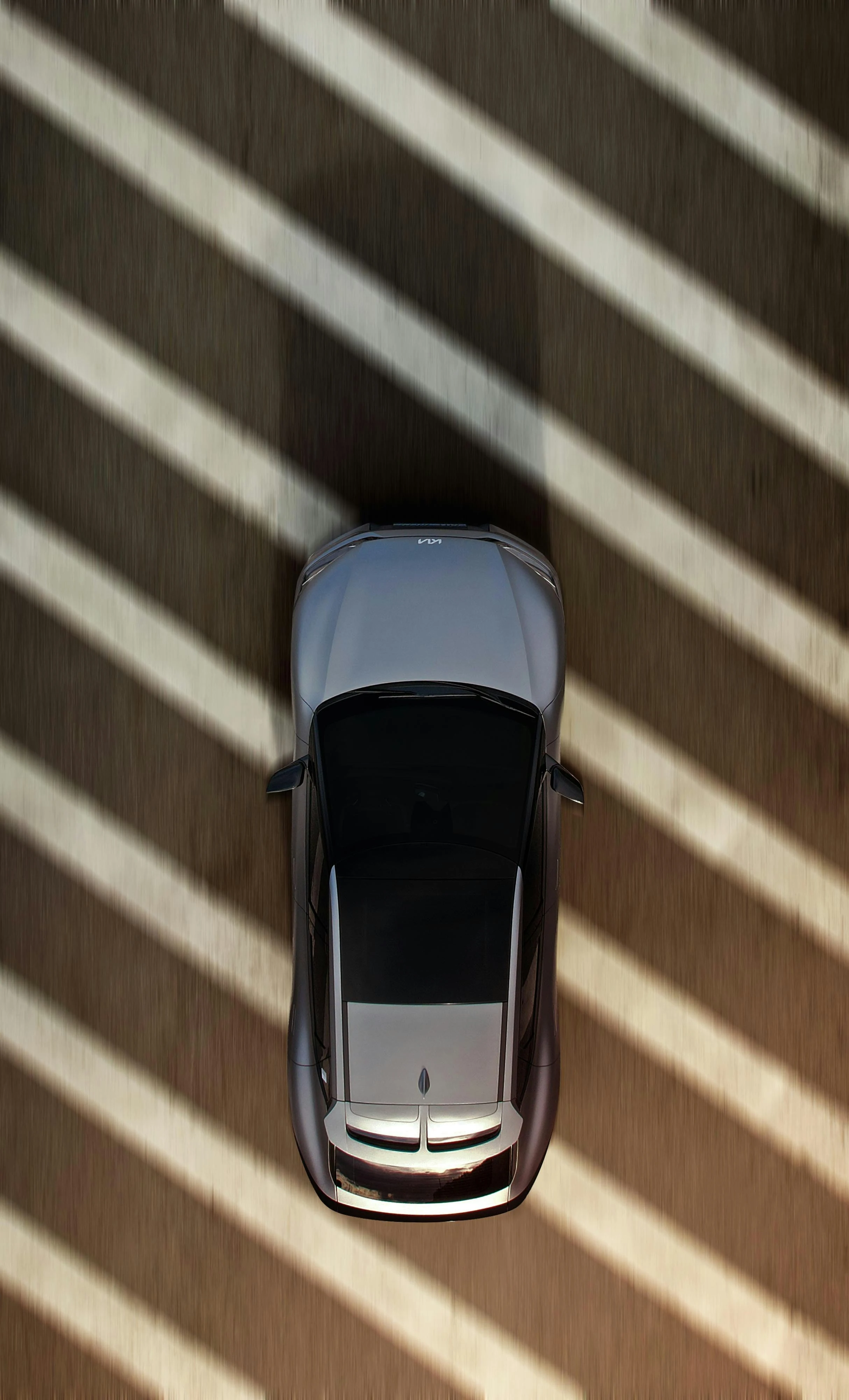Top-down view of a silver electric car driving along a sun-striped road, with diagonal shadows cast across the ground and the vehicle.