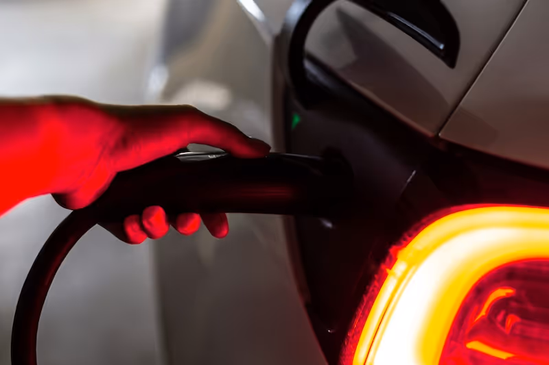 Close-up of a hand plugging a charging cable into an electric car, illuminated by rear brake light.