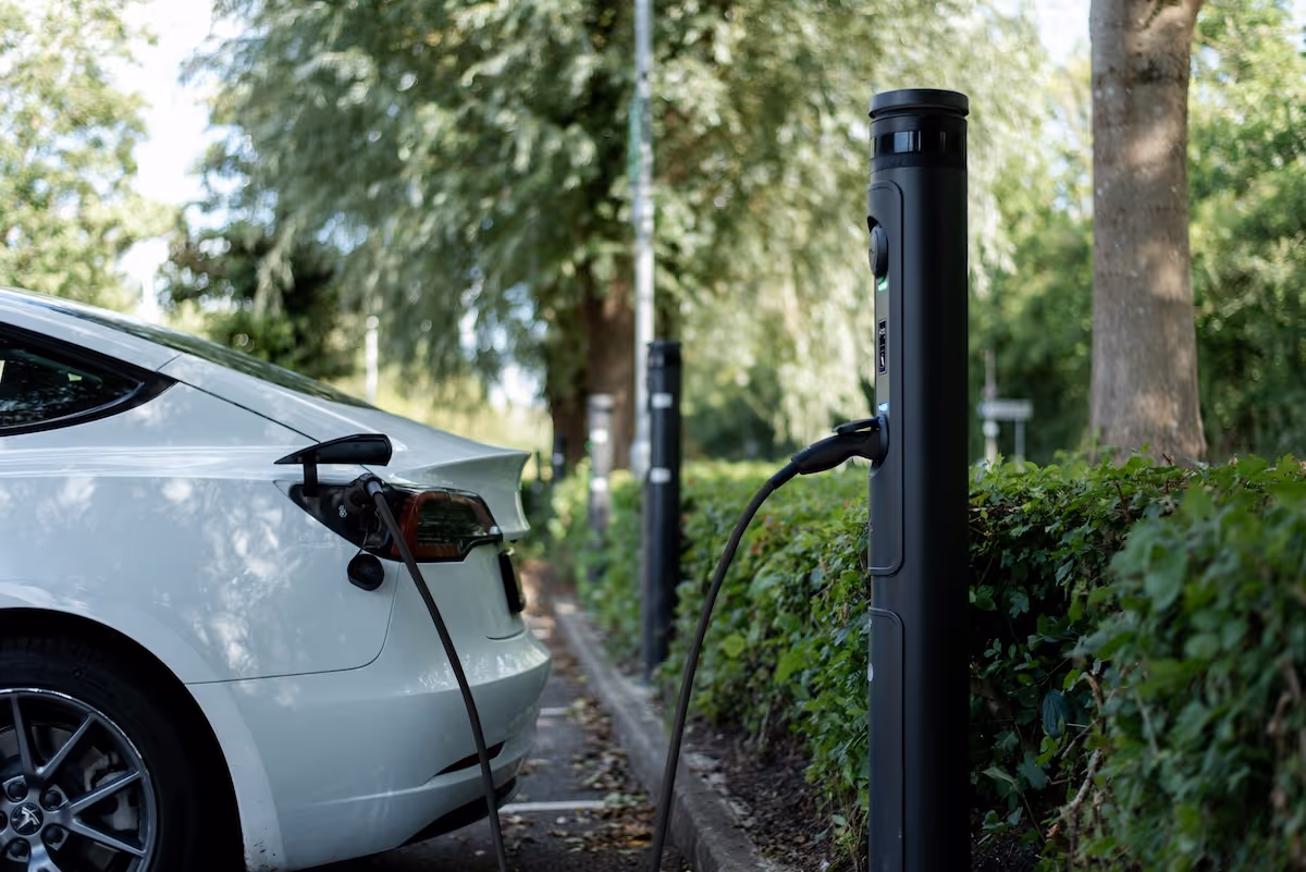 Electric car charging at a public charging post along a tree-lined street.