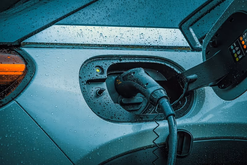 Close-up of an electric car charging port with cable connected, covered in raindrops.