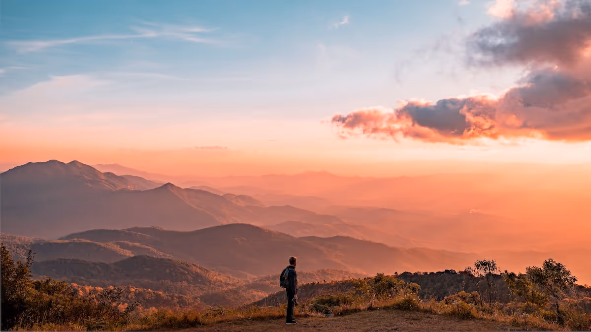 Person standing on a hillside overlooking layered mountain ranges at sunset under a pastel sky.
