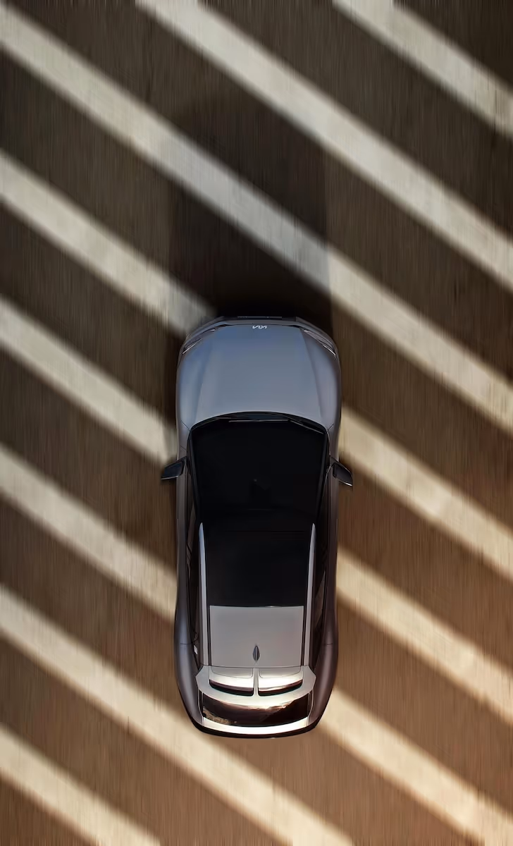 Top-down view of a silver electric car driving along a sun-striped road, with diagonal shadows cast across the ground and the vehicle.