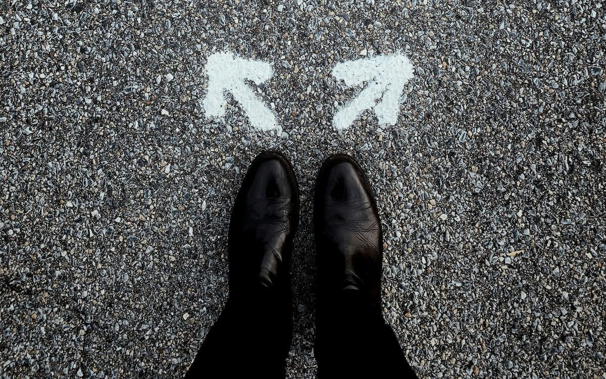 Looking down at a pair of feet with two arrows painted in front of them. One arrow points off to the left, the other to the right, denoting a choice.