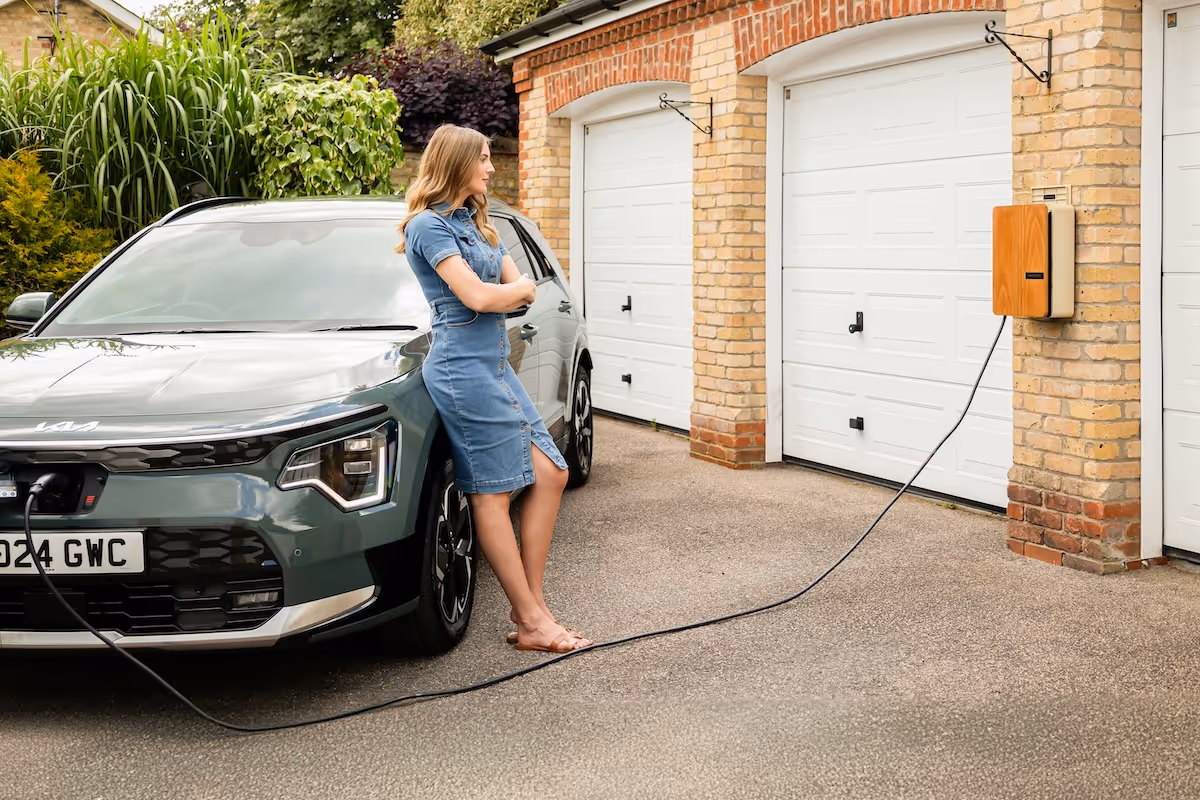 A woman stands against her electric car whilst it's plugged in to a home charger. The charger is installed on a double garage