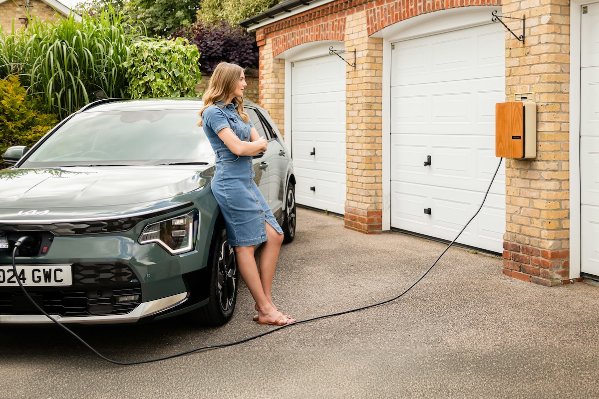 A woman stands against her electric car whilst it's plugged in to a home charger. The charger is installed on a double garage 