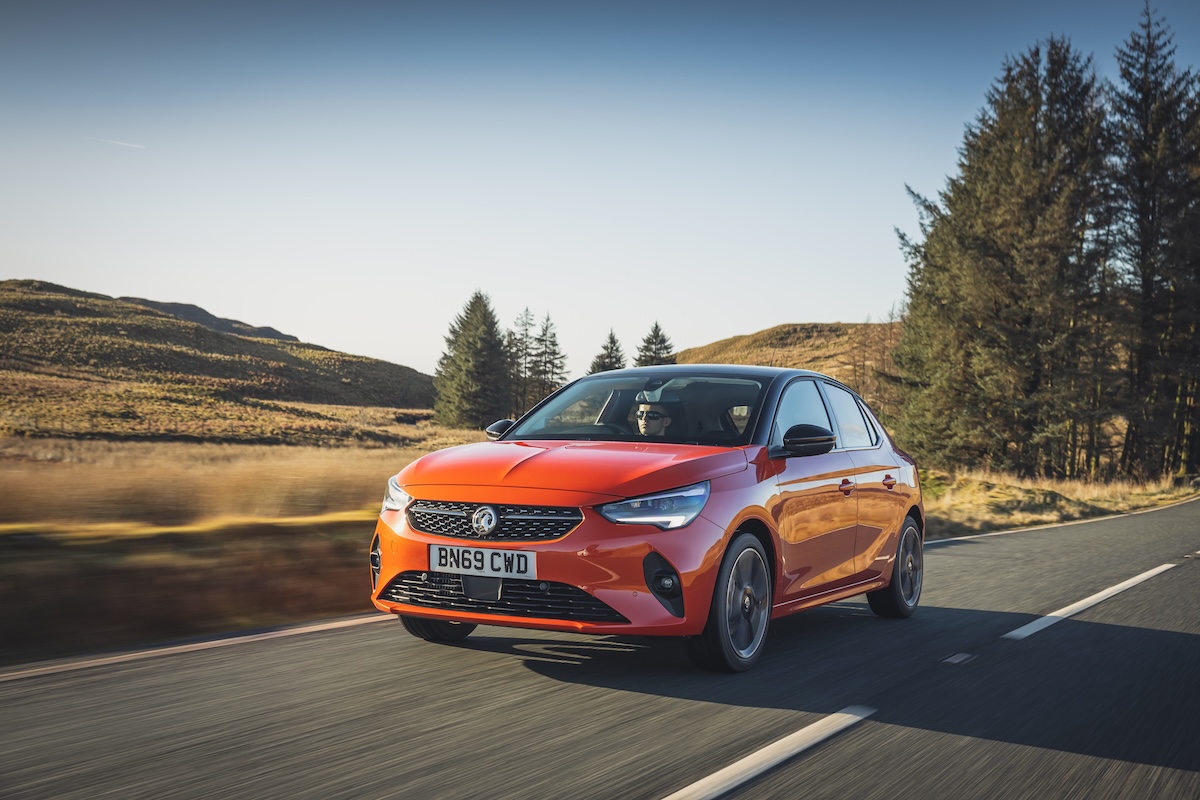 A Vauxhall Corsa electric view from the front. It's orange. Parked on a driveway with a double- garage to the righthand side. 