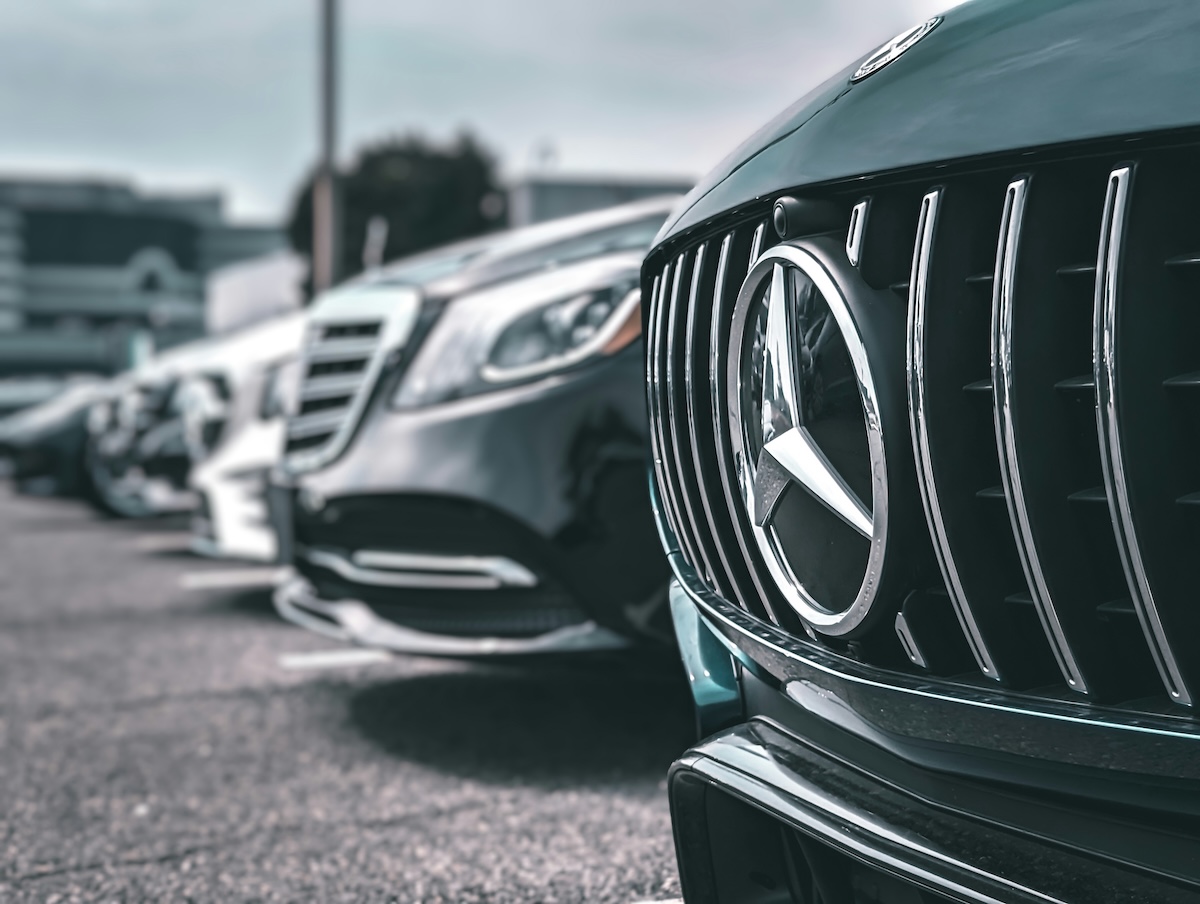 A row of vehicles lined up. The Mercedes-Benz is featured prominently on a black grille next to a selection of other premium vehicles.