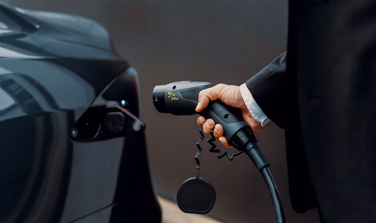 A man's hand holding an EV charging cable, he's dressed in smart wear and is about to plug it into the charging port of a black Tesla 