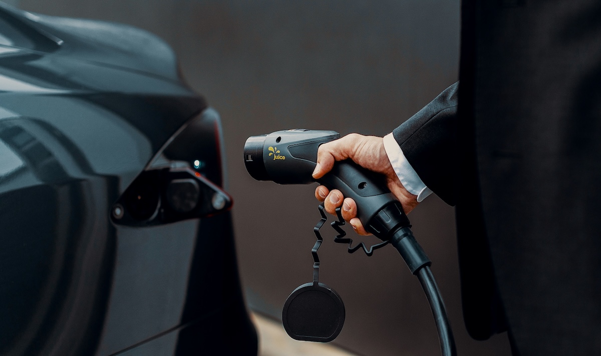 A man's hand holding an EV charging cable, he's dressed in smart wear and is about to plug it into the charging port of a black Tesla