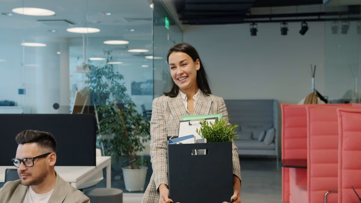 A woman smiles holding a box full of her office equipment, she's walking through an office setting on her way out