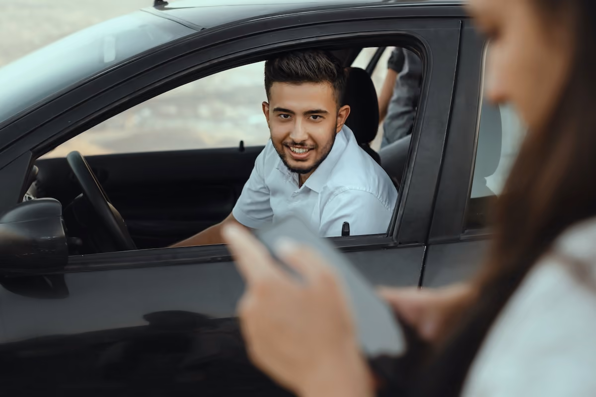 A man smiles out the window of a vehicle, in the foreground a woman holds her phone and looks down at it