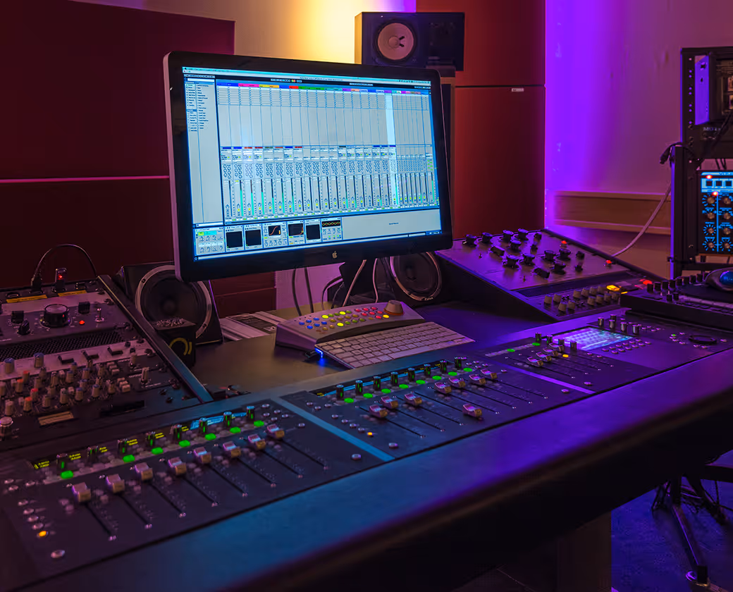 Professional audio mixing console with illuminated controls and a monitor displaying digital audio workstation software in a studio.