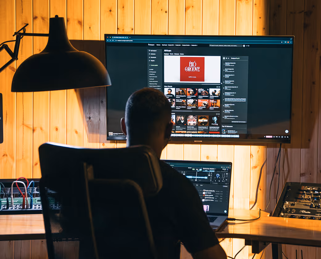 Person sitting in a chair facing two screens on a wooden wall; the larger screen shows a music playlist with an album cover.
