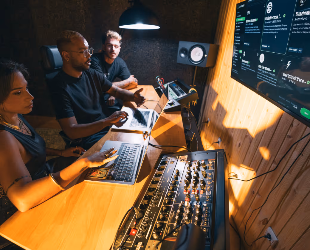 Three people working together in a music studio with laptops, audio mixer, and speaker, discussing music production.