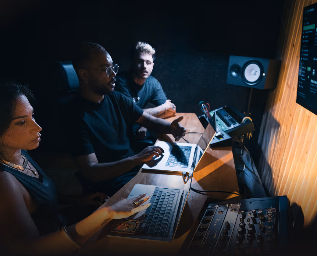 Three people seated in a dimly lit studio working on laptops with audio equipment and a monitor nearby.