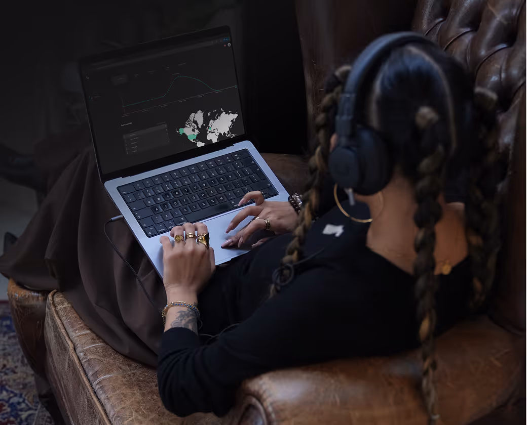 Person with braided hair and rings wearing headphones using a laptop with a data dashboard on screen.