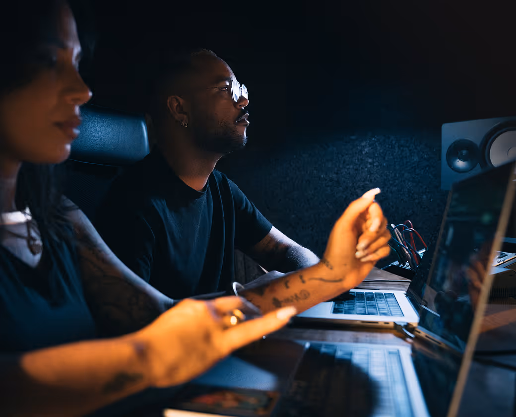 Two people working on laptops in a dimly lit recording studio with audio monitors.