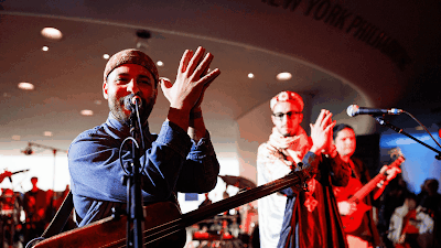 A large, engaged crowd in a three-level hall illuminated by blue lights, watches five musicians perform on stage.