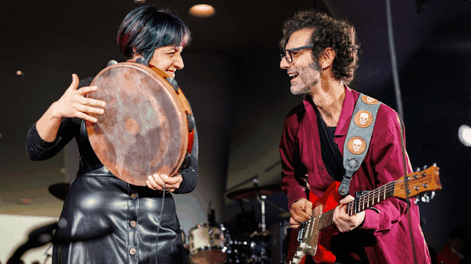 A large, engaged crowd in a three-level hall illuminated by blue lights, watches five musicians perform on stage.