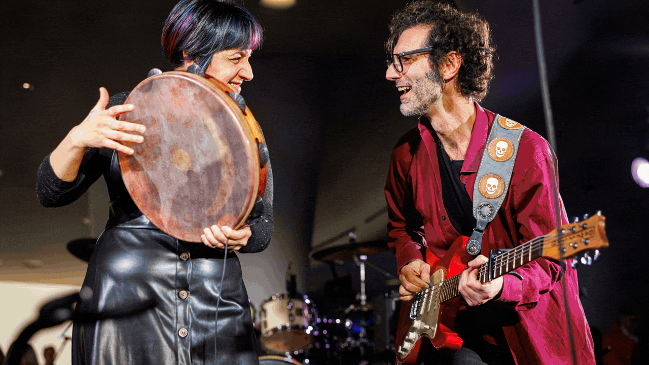 A large, engaged crowd in a three-level hall illuminated by blue lights, watches five musicians perform on stage.