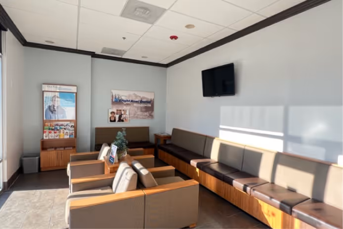 Bright waiting room with brown leather chairs and benches, a wall-mounted TV, and sunlight streaming through windows.