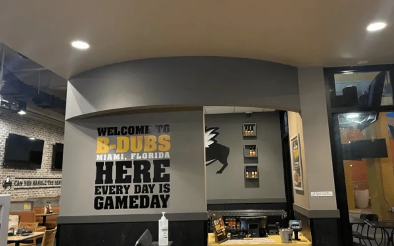 Interior wall of B-Dubs Miami with text reading 'Welcome to B-Dubs Miami, Florida. Here every day is gameday' in black and yellow letters.