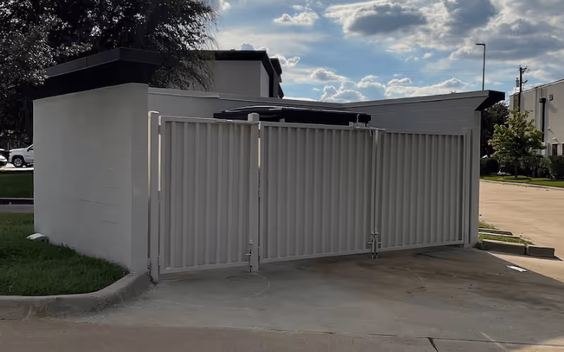 Closed metal gate enclosing an outdoor dumpster area with a concrete wall and buildings in the background under a partly cloudy sky.