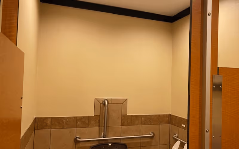 Interior view of a restroom stall with beige tiled walls, metal grab bars, and a toilet seat visible.
