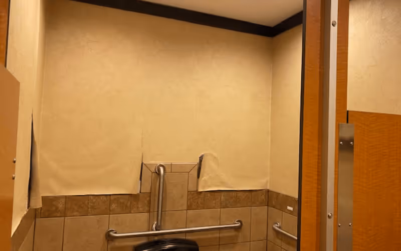 Restroom stall with beige tiled walls, stainless steel grab bars, and a partially covered wall-mounted dispenser.