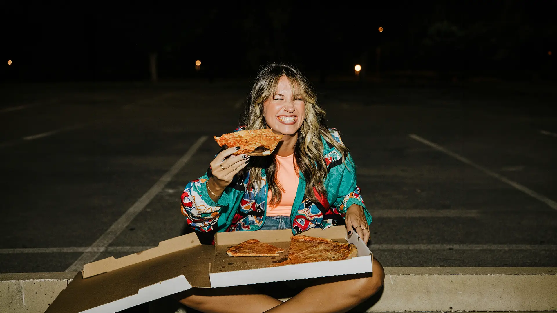 Smiling woman in colorful jacket sitting cross-legged eating a slice of pizza at night in empty parking lot.