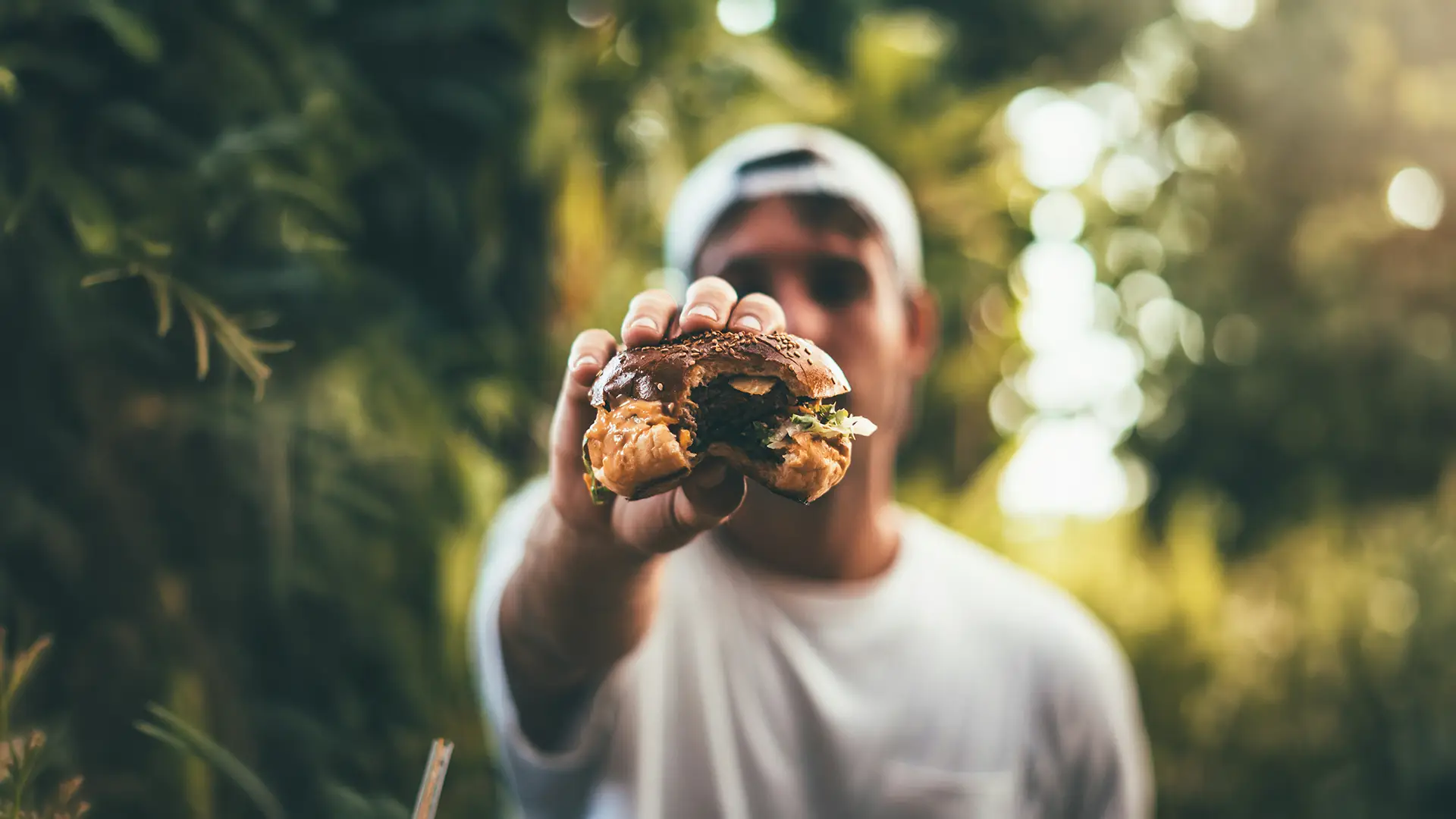 Person holding a bitten cheeseburger toward the camera outdoors with blurred greenery background.