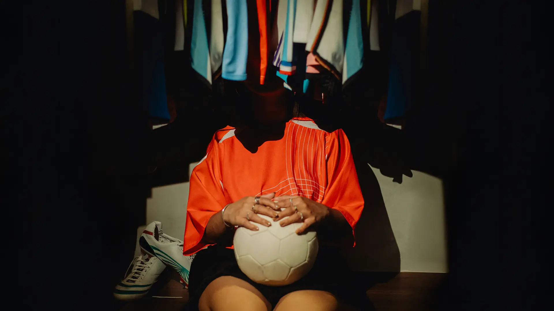Person in an orange jersey holding a soccer ball, sitting on the floor near hanging clothes and cleats.