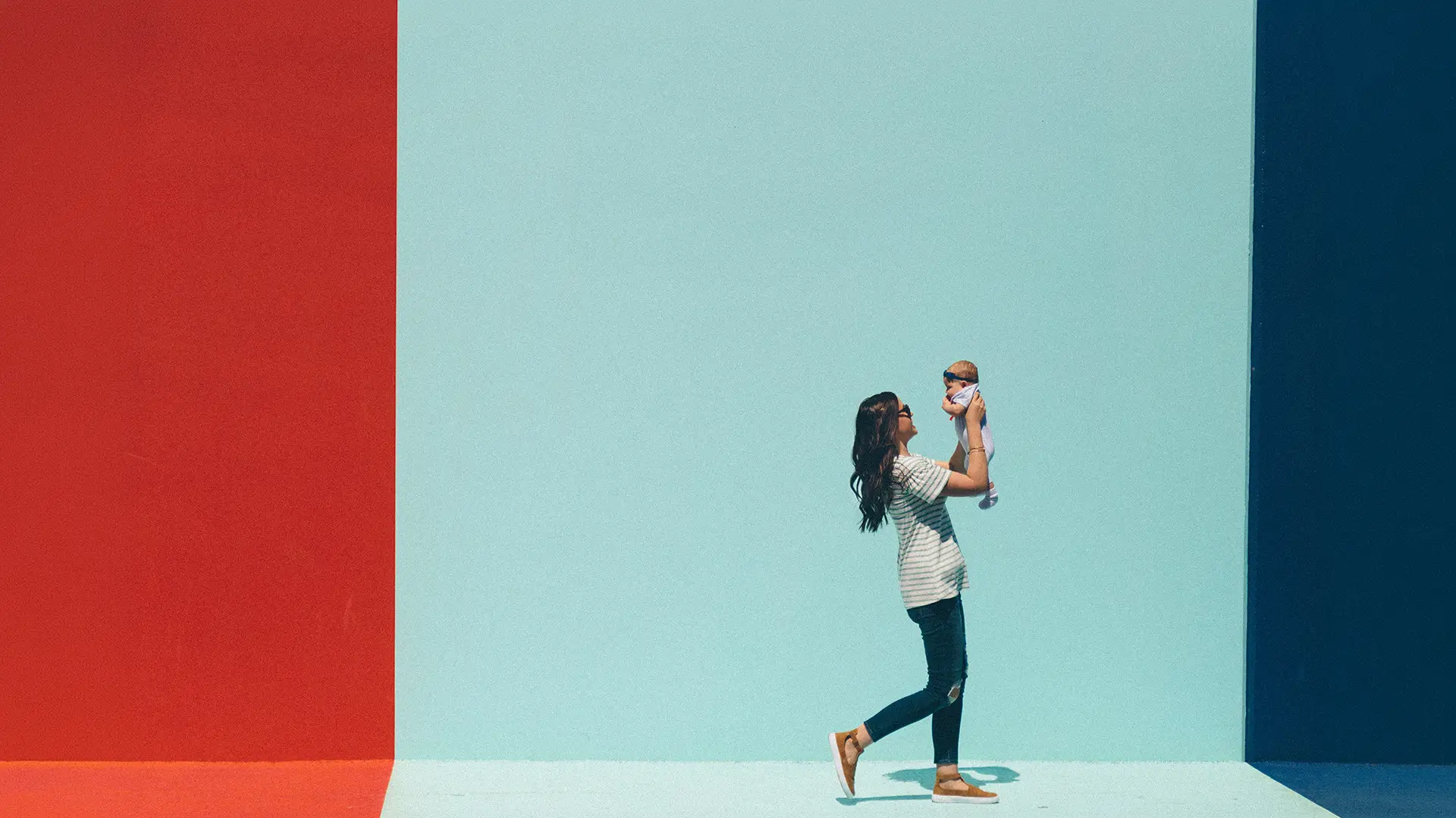 Woman lifting baby in front of a wall divided into red, light blue, and dark blue sections.