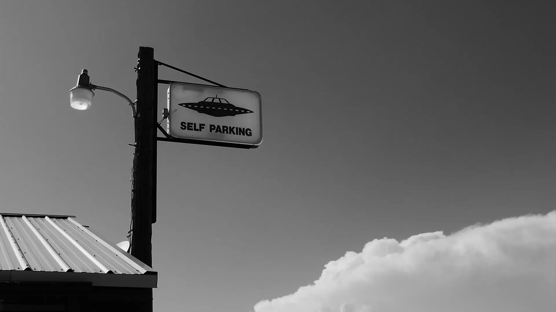 Street lamp pole with a sign showing a UFO and text reading 'SELF PARKING' against sky with clouds.