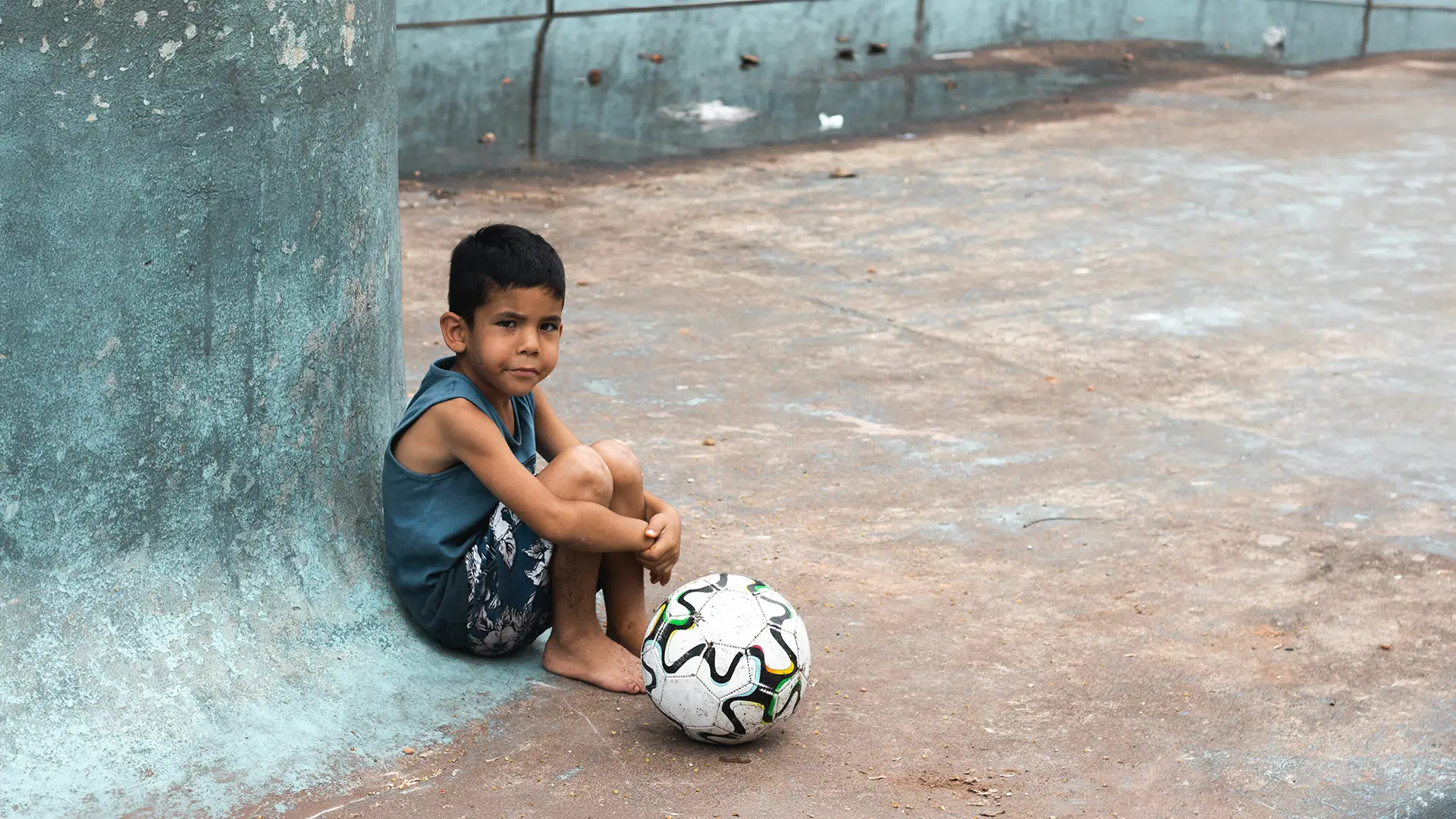 Young boy sitting barefoot on concrete ground next to a curved wall with a soccer ball nearby.