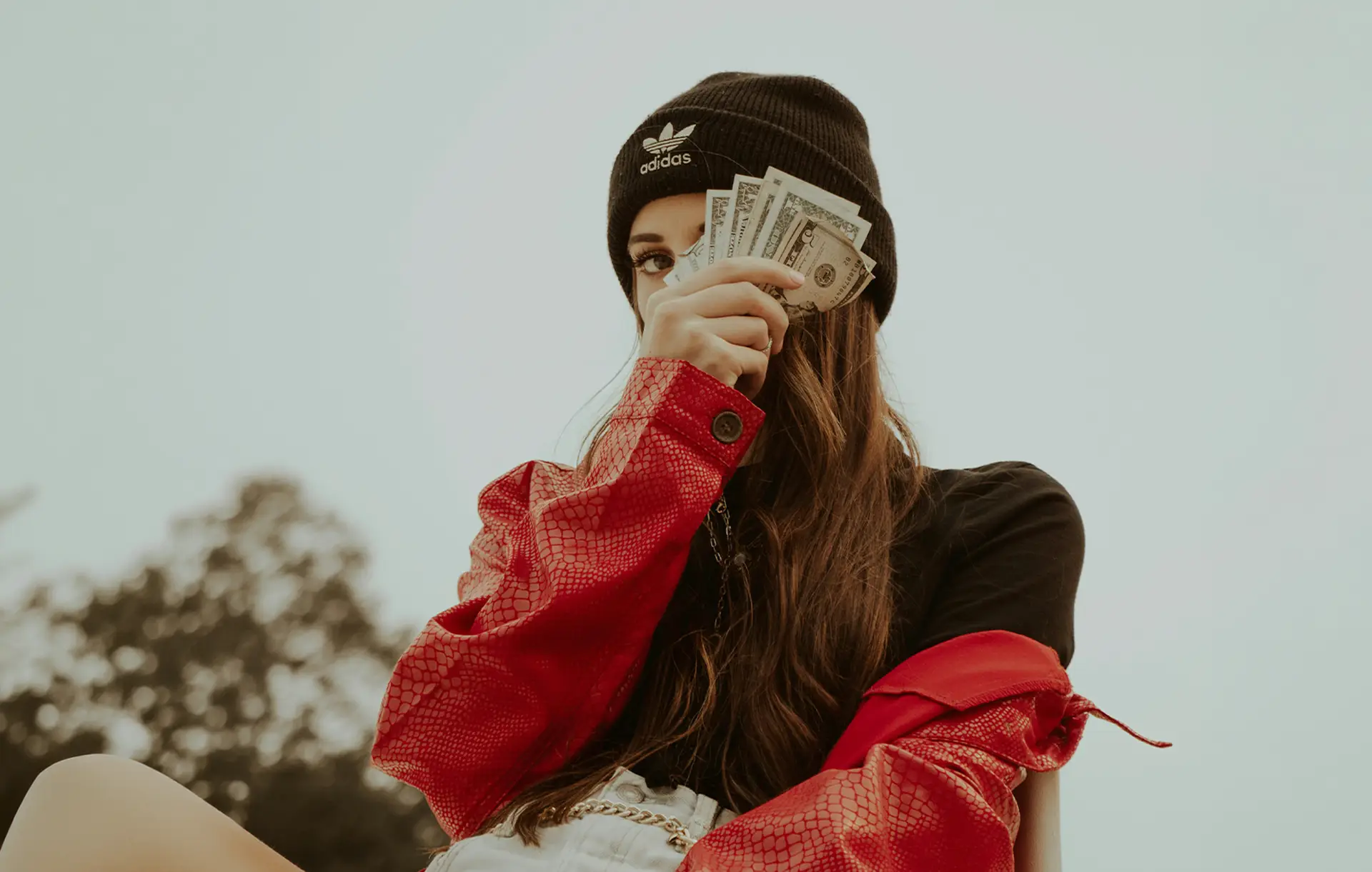 Person in black Adidas beanie and red jacket holding fan of US dollar bills covering part of face.