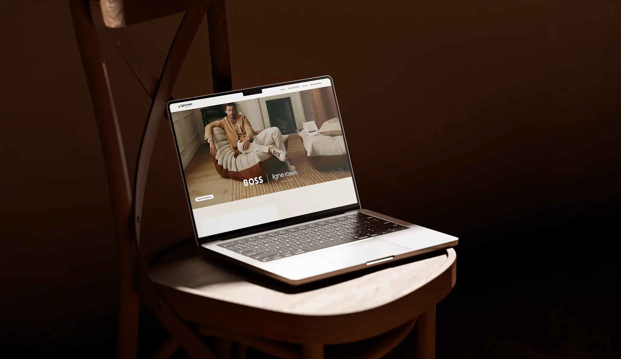 Open laptop displaying a webpage featuring a man sitting on a uniquely designed beige and brown chair, with the text 'BOSS | ligne roset' visible, placed on a wooden chair against a dark brown background.