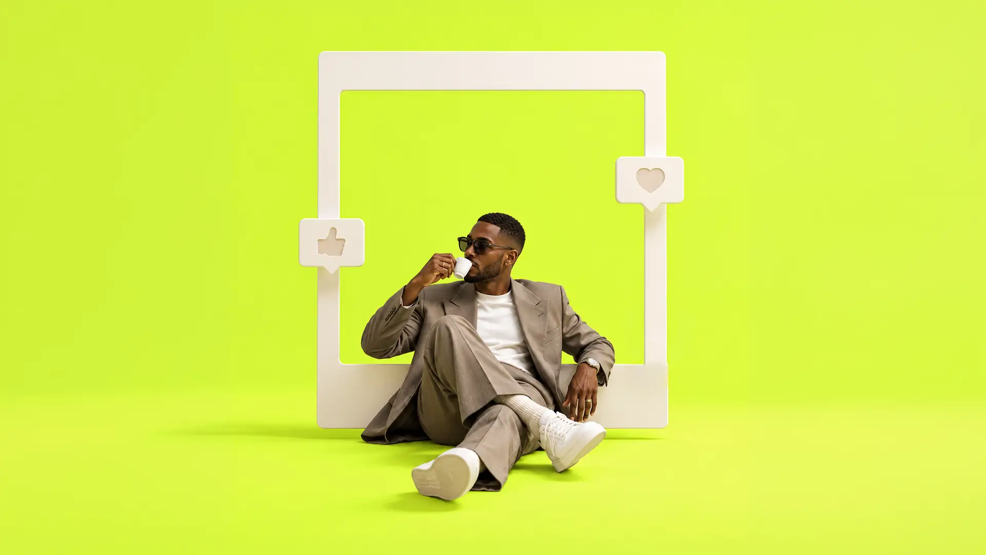 Man dressed in a beige suit, white sneakers, and sunglasses sitting cross-legged on a bright yellow-green floor, sipping from a small white cup, framed by a large white cutout shaped like a social media post with thumbs-up and heart icons on either side.