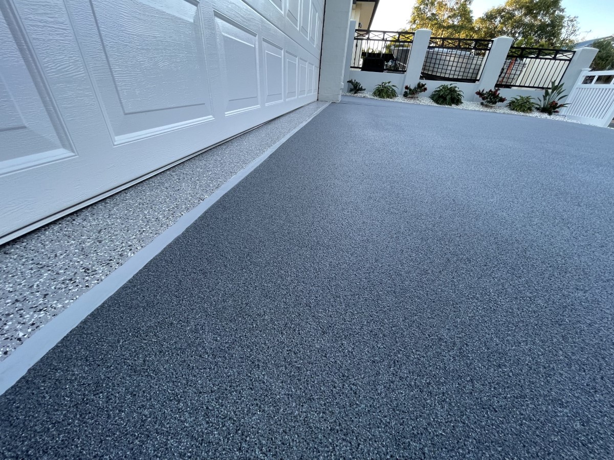 Smooth dark grey driveway floor with speckled border along a white garage door, and a white fence with plants in the background.