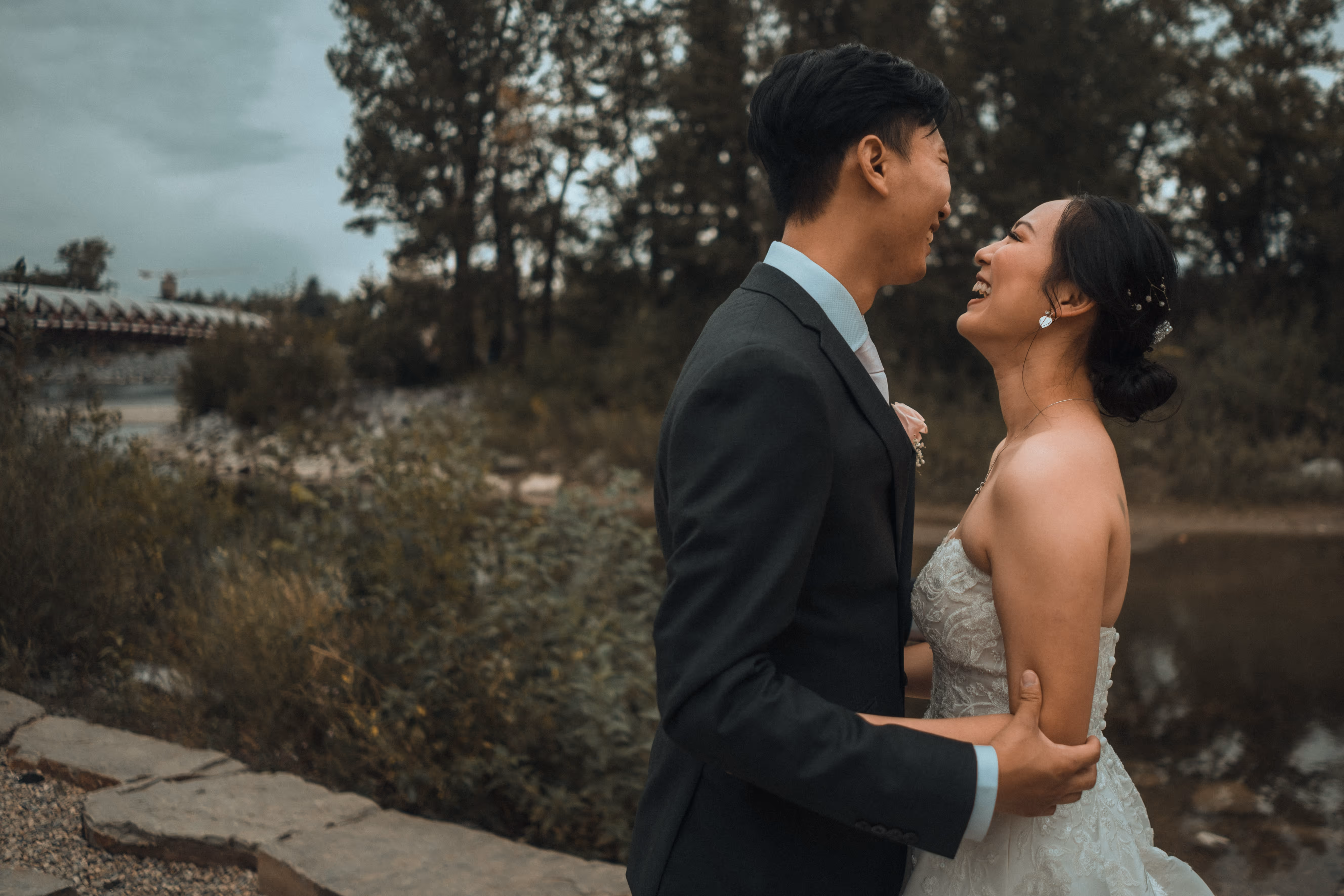 Bride and groom laughing and embracing outdoors near a river and trees.