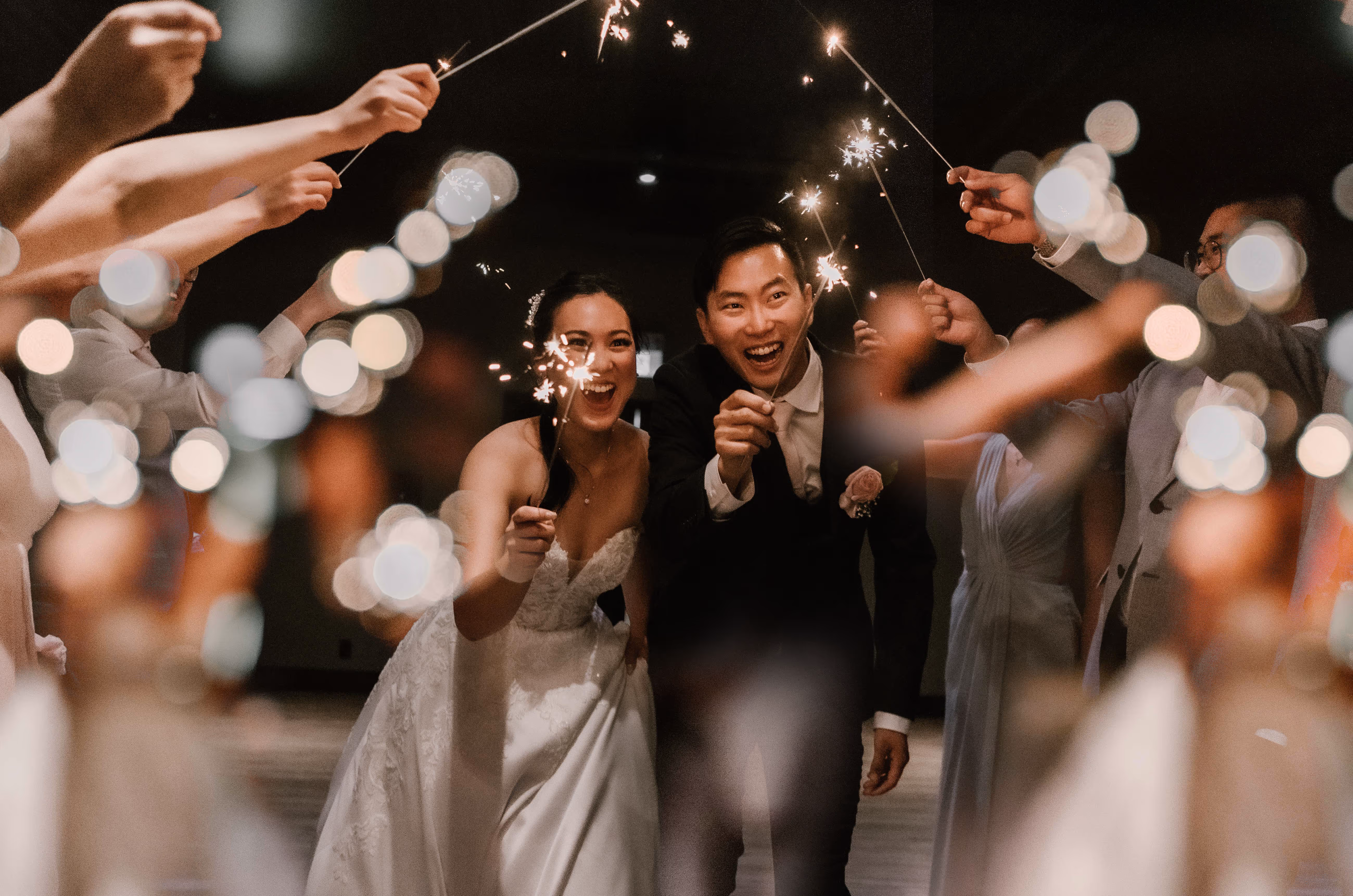 Bride and groom smiling and holding sparklers surrounded by guests also holding sparklers at a wedding celebration.