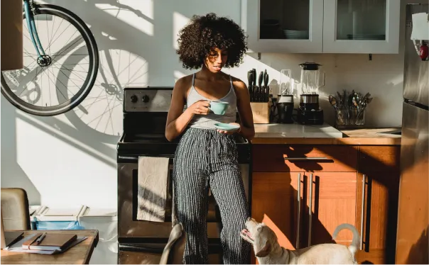 Woman drinking a cup of tea in her kitchen looking at pet dog